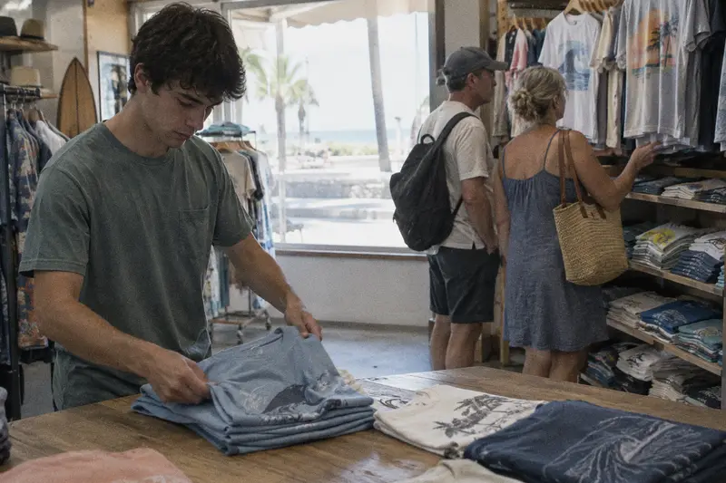 Un dependiente dobla camisetas mientras una pareja de turistas mira la ropa al fondo de una tienda de playa