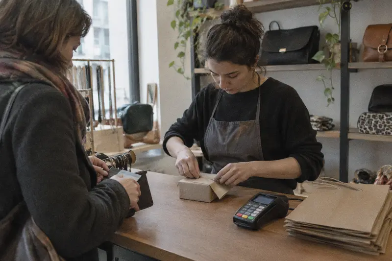 Una dependienta envuelve un regalo en el mostrador mientras la clienta guarda la tarjeta después de pagar