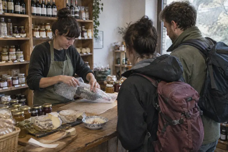 Una dependienta envuelve una botella de vino para el viaje mientras una pareja con mochilas espera en el mostrador