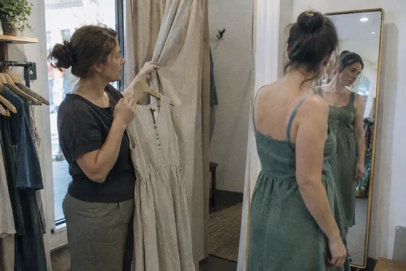 Una dependienta ofrece otra talla de un vestido junto al probador mientras la clienta se mira en el espejo