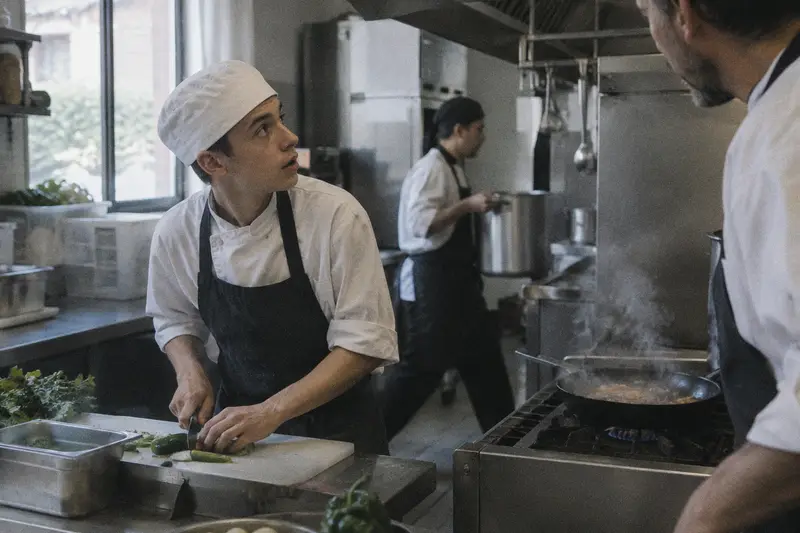 Un ayudante de cocina contesta a su jefe mientras un compañero pasa por detrás con una olla
