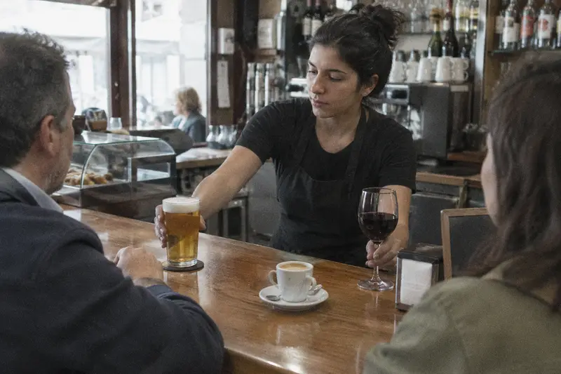 Una camarera sirve una caña y un tinto a dos clientes en la barra de un bar