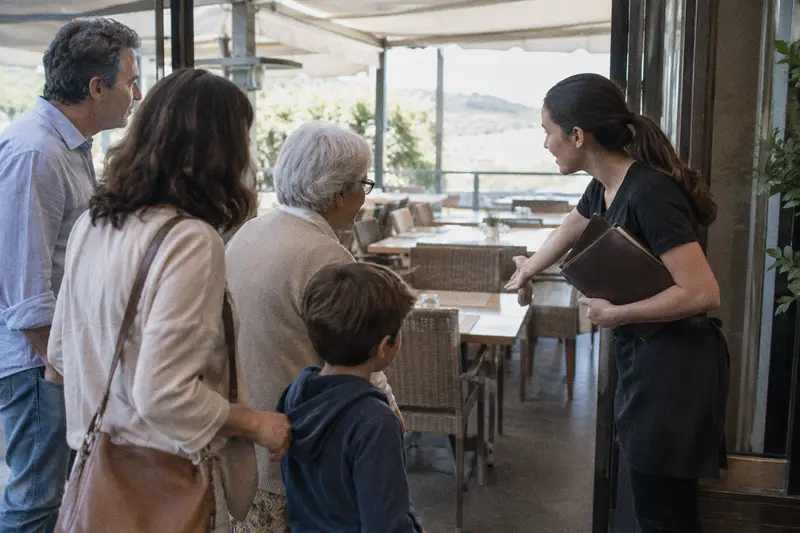 Una camarera recibe a una familia en la terraza de un restaurante y les señala una mesa libre