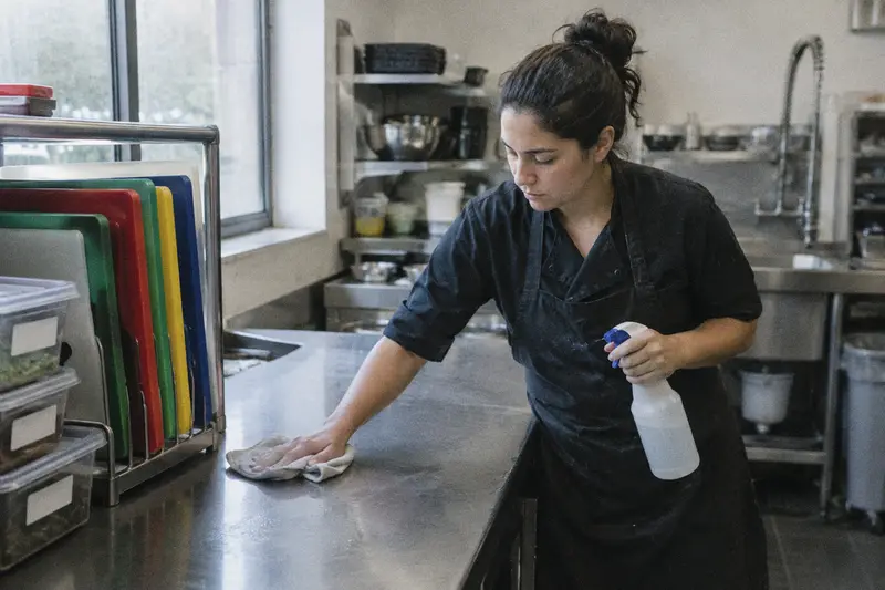 Una ayudante de cocina limpia su mesa de acero junto a un soporte con tablas de colores