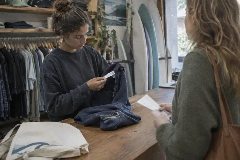 Una dependienta revisa la etiqueta de una sudadera que una clienta trae para cambiar