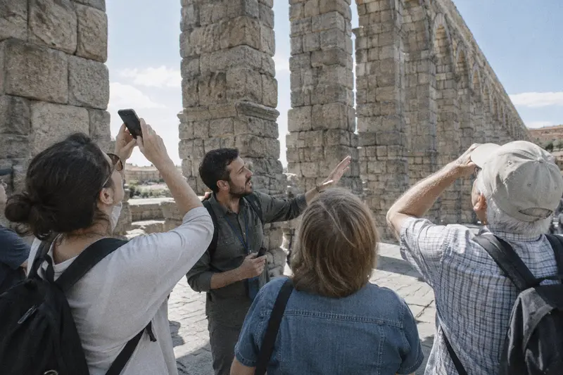 Un guía turístico explica un acueducto romano de piedra a un grupo de turistas que miran hacia los arcos