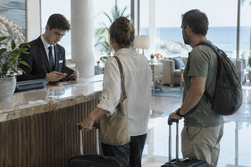 Un recepcionista revisa un pasaporte mientras una pareja de turistas espera con sus maletas en el mostrador