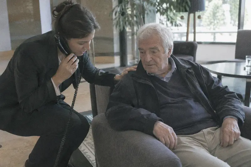 Una recepcionista acompaña a un huésped mayor sentado en el vestíbulo mientras llama por teléfono
