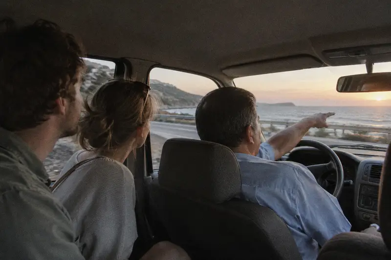 Un taxista señala hacia el mar a una pareja de turistas que miran por la ventanilla en una carretera de costa