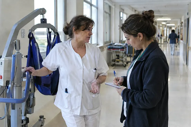 Una celadora habla con la delegada de prevención en el pasillo de un hospital, junto a una grúa de pacientes